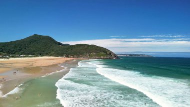 Berrak mavi gökyüzünün altında dalgaların, kumlu kıyıların ve yemyeşil yeşilliklerin olduğu çarpıcı bir sahil manzarası..