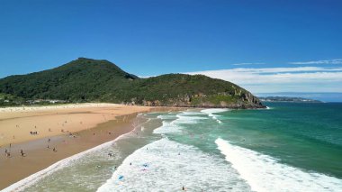 Güzel bir sahil, altın kumlar üzerinde yumuşak dalgalar, yemyeşil tepeler ve yukarıda parlak mavi bir gökyüzü.