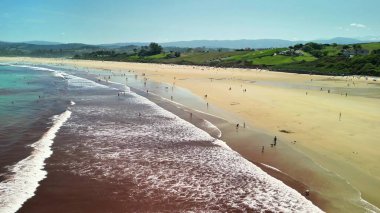 Bu hava fotoğrafında dinamik dalgalar, güneş banyosu ve neşe uyandıran nefes kesici manzaralarla dolu canlı bir plaj var.