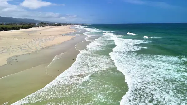 Sakin ve davetkar bir plaj sahnesi yumuşak dalgalar, güzel altın kum ve parlak mavi gökyüzü.