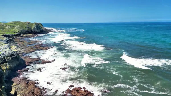 Çarpıcı hava görüntüleri engebeli uçurumlara çarpan dinamik dalgalarla dolu canlı bir kıyı manzarasını vurguluyor.