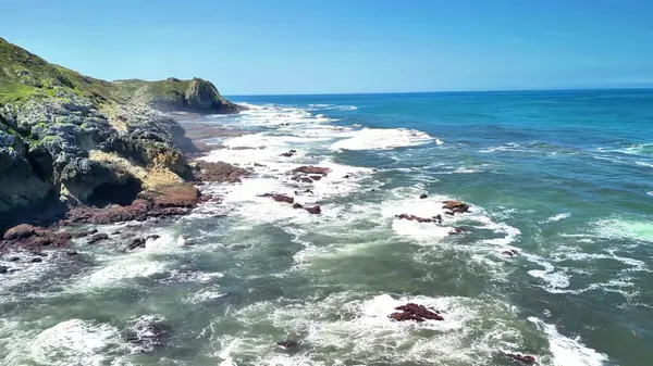Görkemli okyanus dalgalarının açık mavi gökyüzünün altında kayalık kıyılara çarptığını gösteren nefes kesici bir sahne.