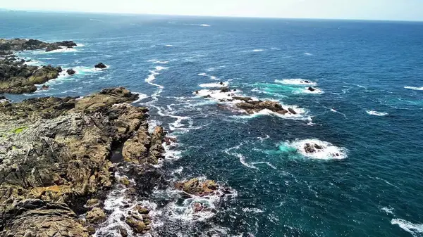 Turkuaz deniz dalgalarıyla birleşen kayalık bir sahil şeridinin çarpıcı hava görüntülerini görkemli bir manzara için tecrübe edin.