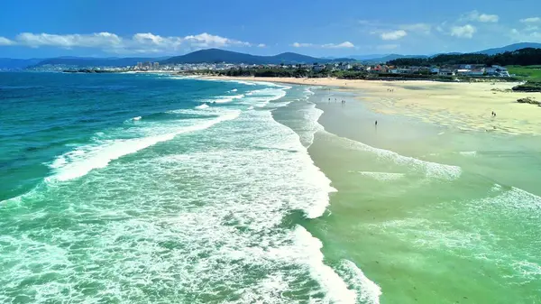 Hava fotoğrafı, berrak mavi gökyüzünün altında sakin sahil dalgalarının ve sahil şeridinin nefes kesici görüntülerini yakalıyor.