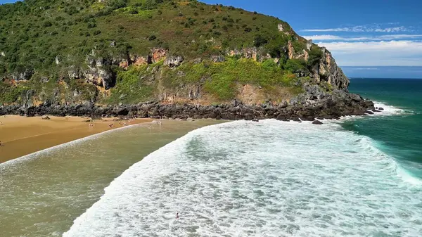 Dalgaların güçlü bir şekilde yemyeşil kayalıkların altındaki kumlu bir sahile çarptığını hissedin.