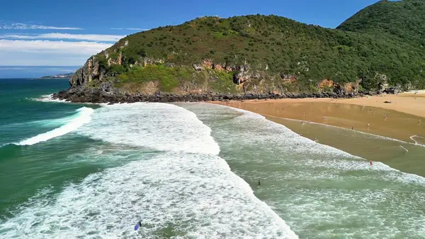 Yumuşak, dalgalı, yemyeşil tepeler ve berrak mavi gökyüzü içeren huzurlu bir kumsalın hava görüntüsü.