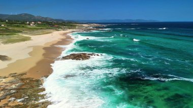 Berrak turkuaz suları ve yumuşak dalgaları olan sakin bir plajın güzelliğini gösteren bir hava çekimi..