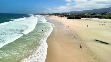Nefes kesen bir kıyı manzarası. Dalgalar yumuşak kuma çarpıyor. İnsanlar kumsalın tadını çıkarıyor..