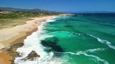 Turkuaz suları, altın kumları ve kıyıya vuran dalgalarla nefes kesici bir sahilin havadan görüntüsü.