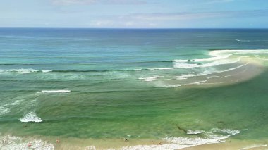 Zarif okyanus dalgalarının büyüleyici görüntüsü berrak mavi gökyüzünün altında yumuşak, kumlu plaja doğru nazikçe süzülüyor.