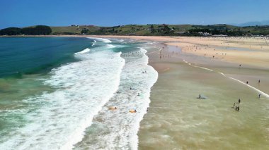 Güneşli bir günde dalgaların, güneşlenenlerin ve açık mavi gökyüzünün sergilendiği güzel bir sahil manzarası.
