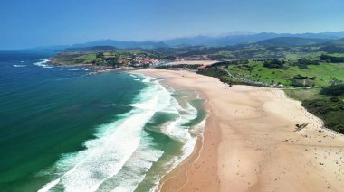 Dalgalar, altın kumlar ve mavi gökyüzünün altında yemyeşil bir kumsalın çarpıcı manzarası.