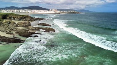 Kıyı şeridinin çarpıcı manzarası, dalgaların çarpması ve arka planda bir şehir. Seyahat ve doğa temaları için ideal.