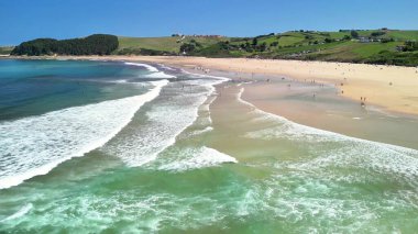 Hareketli dalgaların, geniş kumlu kıyıların ve parlak mavi gökyüzünün altındaki yemyeşil tepelerin yer aldığı huzurlu bir sahil sahnesi.