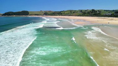 Bu çarpıcı hava manzarası, berrak mavi gökyüzünün altında sakin bir kıyı şeridine çarpan canlı dalgaları canlı bir şekilde gösteriyor.