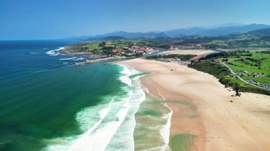 Arka planda turkuaz suları ve yemyeşil tepeleri olan çarpıcı bir plajı gösteren hava fotoğrafı.