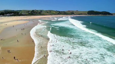 Zarif dalgalar, parlak güneş ışığı ve sıcak güneşin keyfini çıkaran ziyaretçilerle çarpıcı bir sahil manzarası.