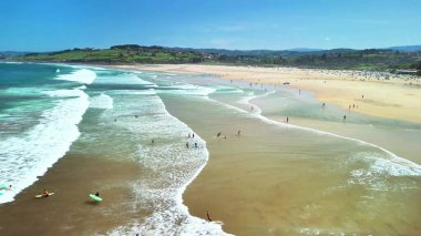 Güzel, canlı, çarpıcı dalgalar, güneş banyosu ve yemyeşil tepeler sergilenen bir plaj sahnesi.