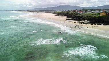 Hava manzarası dalgalı, kayalık kıyıları olan güzel bir sahil ve sıcak, güneşli bir günün tadını çıkaran insanlar gösteriyor.