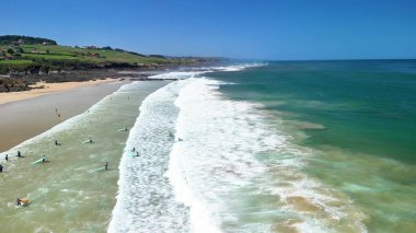 Sörfçülerin, çarpan dalgaların ve açık gökyüzünün altındaki yemyeşil tepelerin olduğu güzel bir kumsal sahnesi yaşayın.