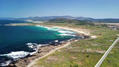 Hava manzarası turkuaz suları, yeşil tarlaları ve açık mavi gökyüzü olan güzel bir kıyı şeridini gösteriyor.