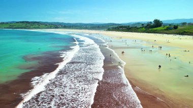 Güneşin tadını çıkaran ve sakin bir sahilde sörf yapan ziyaretçileri gösteren nefes kesici bir sahil manzarası.