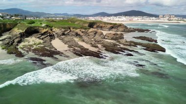 Kayalık bir sahilin nefes kesici manzarası, kıyıya vuran dalgalar ve arka planda manzaralı tepeler..