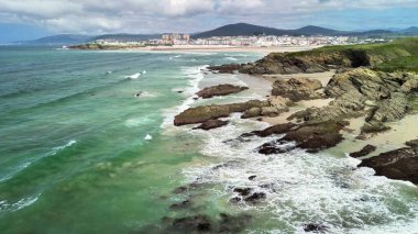 Güzel okyanus dalgaları uzak binaları ve tepeleri olan sakin bir sahil boyunca kayalara çarpıyor..