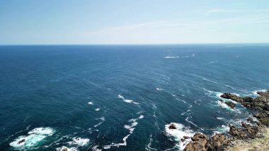 Yüksek bir uçurumdan nefes kesen okyanus manzarası güzel bir şekilde dalgaların mavi gökyüzünün altında yuvarlandığını gösteriyor.