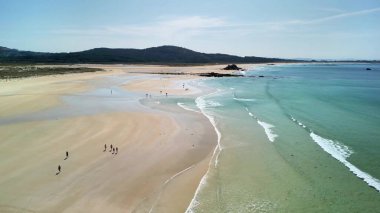 Kumlu sahil boyunca rahatça yürüyen ve dalgaların nazikçe dalgalanmasının tadını çıkaran insanların olduğu pitoresk bir sahil sahnesi.