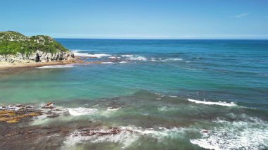 Berrak turkuaz suların ve parlak mavi gökyüzünün altında engebeli kıyı şeritlerinin yer aldığı sakin ve pitoresk bir sahil manzarası.