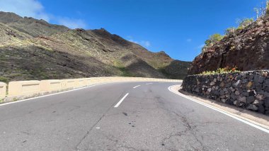 Manzaralı bir kıyı yolu görkemli tepeler boyunca zarif bir şekilde esiyor ve üzerinde parlak mavi bir gökyüzü olan yüksek dağlar.