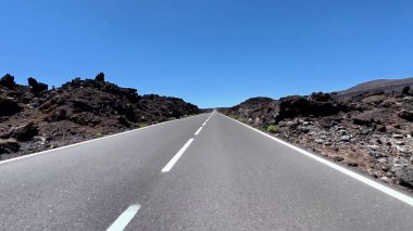 Açık mavi gökyüzü altında volkanik kaya oluşumları boyunca uzanan düz bir yolun çarpıcı görüntüsü büyüleyici.
