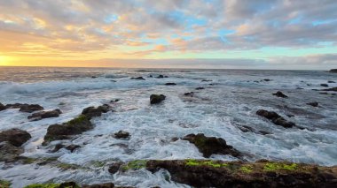 Güzel, sakin bir günbatımı manzarası kayalık kıyıların üzerinde, yumuşak dalgalar çakıyor, çarpıcı güzellikler sergiliyor.