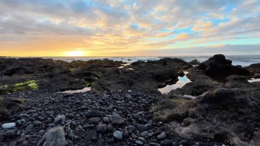 Göz kamaştırıcı bir günbatımı manzarası etraflarındaki kayalık sahili ve sakin suları parlak bir şekilde aydınlatıyor.