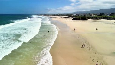 Nefes kesen bir kıyı manzarası. Dalgalar yumuşak kuma çarpıyor. İnsanlar kumsalın tadını çıkarıyor..
