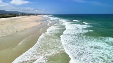 Sakin ve davetkar bir plaj sahnesi yumuşak dalgalar, güzel altın kum ve parlak mavi gökyüzü.