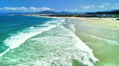 Hava fotoğrafı, berrak mavi gökyüzünün altında sakin sahil dalgalarının ve sahil şeridinin nefes kesici görüntülerini yakalıyor.