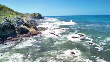 Görkemli okyanus dalgalarının açık mavi gökyüzünün altında kayalık kıyılara çarptığını gösteren nefes kesici bir sahne.