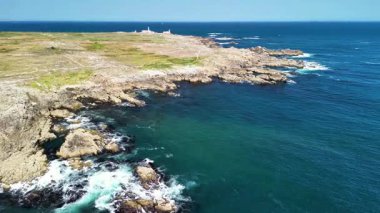 Çarpıcı hava manzaraları, parlak mavi gökyüzünün altında kristal berrak okyanus sularıyla buluşan dramatik kayalık bir kıyı şeridini vurguluyor.