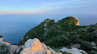 Serin denize bakan kayalık bir tepeden çarpıcı bir manzara, yemyeşil, canlı bitki örtüsüyle çevrili.