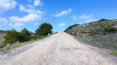 Güzel, manzaralı çakıllı bir yol, parlak, yemyeşil ve kayalık bir arazide, berrak mavi gökyüzünün altında yol alır.