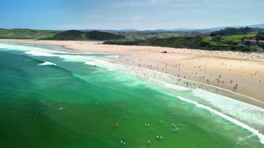 Sersemletici bir hava görüntüsü canlı bir plaj sahnesi, açık gökyüzünün altında sörfçülerle ve güneşlenenlerle dolu..