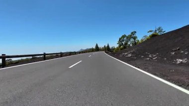 Parlak mavi gökyüzünün altında yemyeşil ağaçlar ve eşsiz lav oluşumları arasında güzel bir şekilde kıvrılan pitoresk dağ yolu.