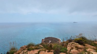 Güzel ve sakin bir kıyı manzarası kayalık kayalıklar, durgun masmavi sular ve bulutlu bir gökyüzü.