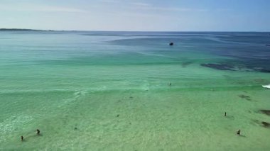 Sakin bir sahil manzarası berrak turkuaz sular, suyun tadını çıkaran insanlar ve uzak yelkenliler..