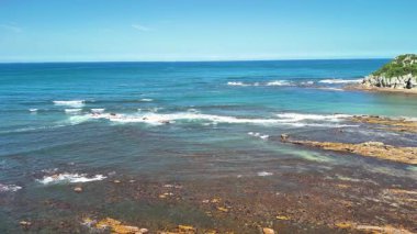 Sarsılan dalgaları, engebeli kayalık kıyı şeridini ve parlak mavi gökyüzünü gösteren güzel, sakin bir okyanus manzarası.
