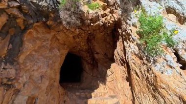 Etrafı engebeli, kayalık arazi ve yemyeşil yemyeşil ile çevrili karanlık bir mağara girişi büyüleyici bir manzara.
