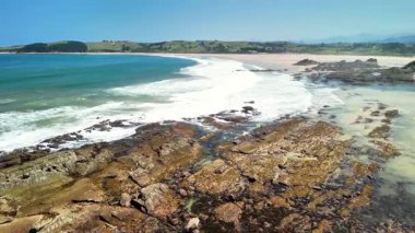 Nefes kesen güzellikte bir kıyı manzarası. Açık mavi gökyüzünün altındaki engebeli kayalara vuran dalgalar.