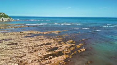 Parlak güneşli bir gökyüzü altında kayalık bir kıyı şeridi ve berrak okyanus dalgalarının çarpıcı görüntüsü huşu uyandırıyor.